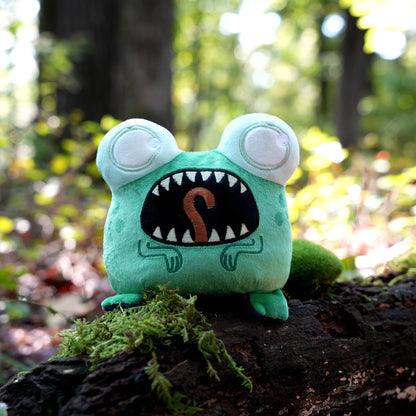 Twisted Cryptids: Loveland Frog 4" Reversible Plushie screaming side sitting on a mossy log in a forest